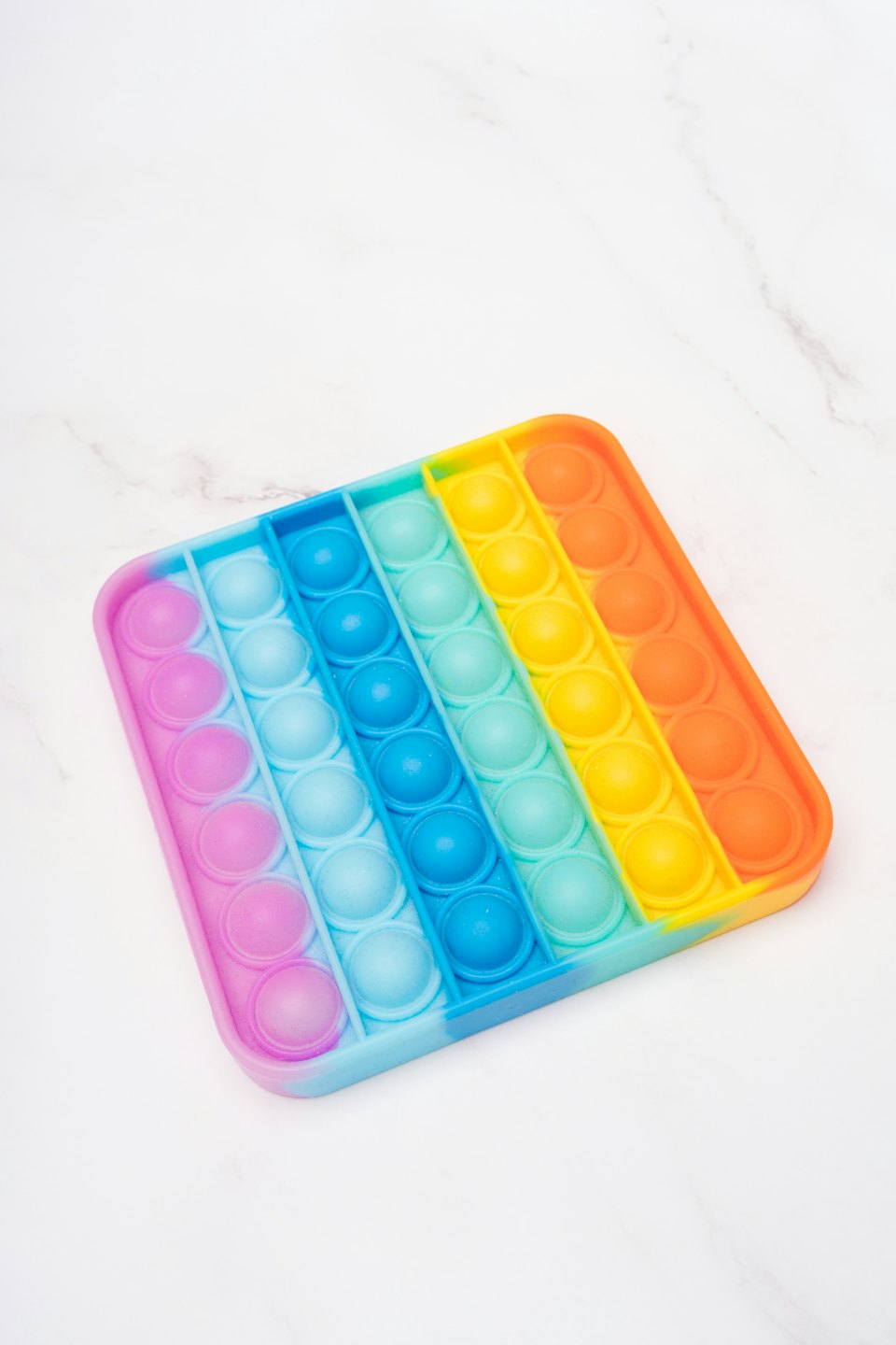 A rainbow colored square pop-it toy sits on white marble.
